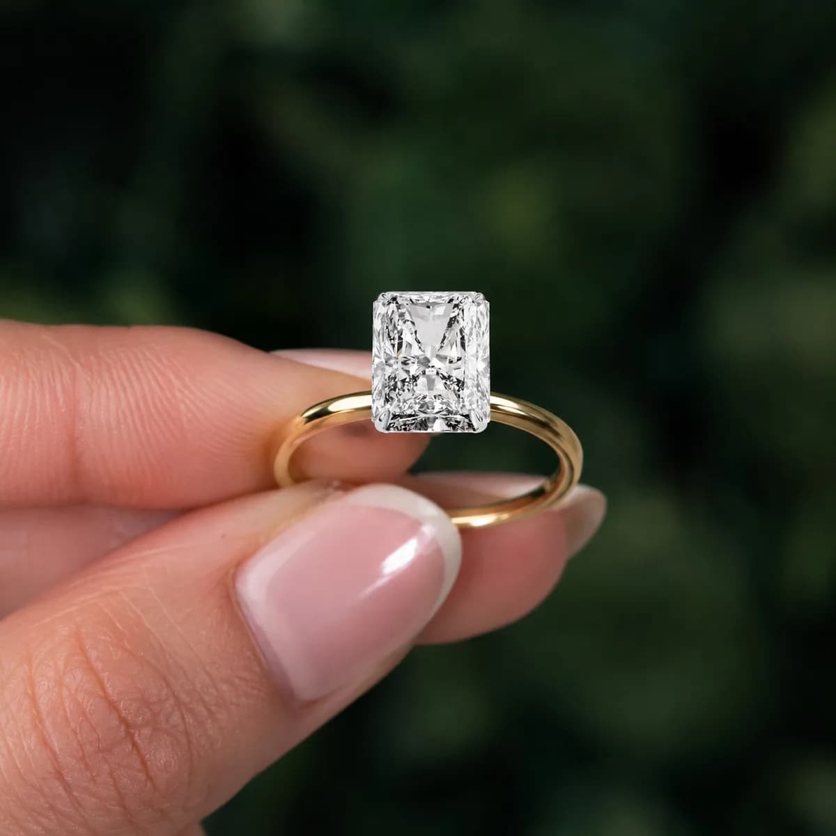 Radiant cut lab grown diamond four-claw solitaire engagement ring in yellow gold held between fingertips