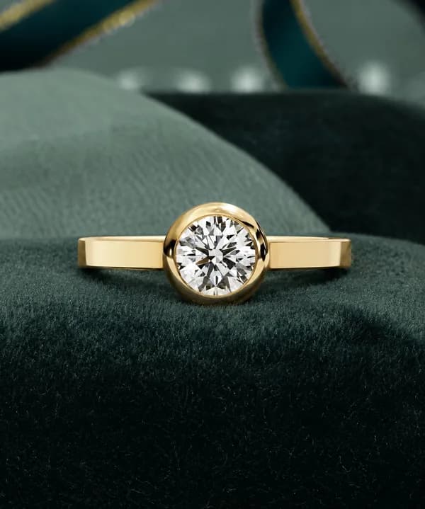 Round lab grown diamond bezel set solitaire engagement ring in yellow gold resting on a green velvet cushion