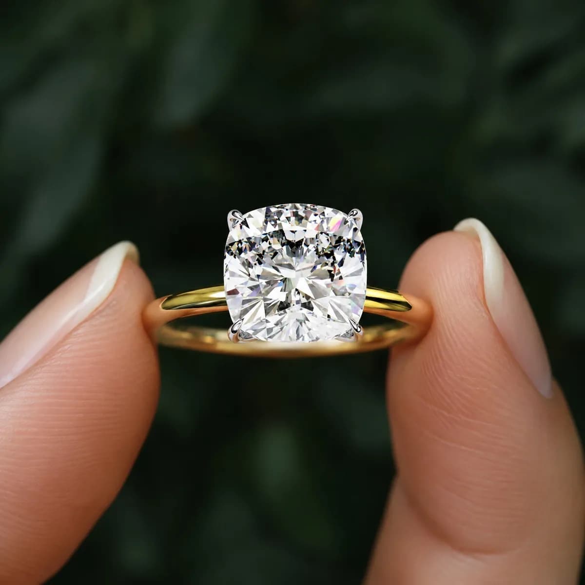 Cushion cut lab grown diamond four-claw solitaire engagement ring in yellow gold held between fingertips