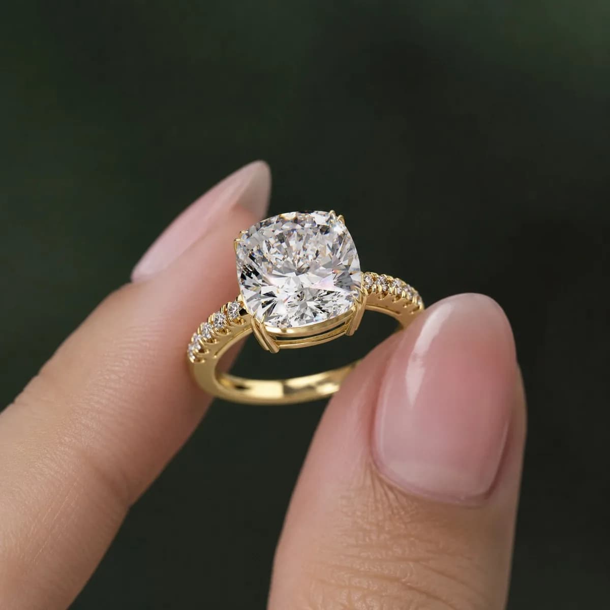 Cushion cut lab grown diamond double claw pavé engagement ring in yellow gold held between fingertips