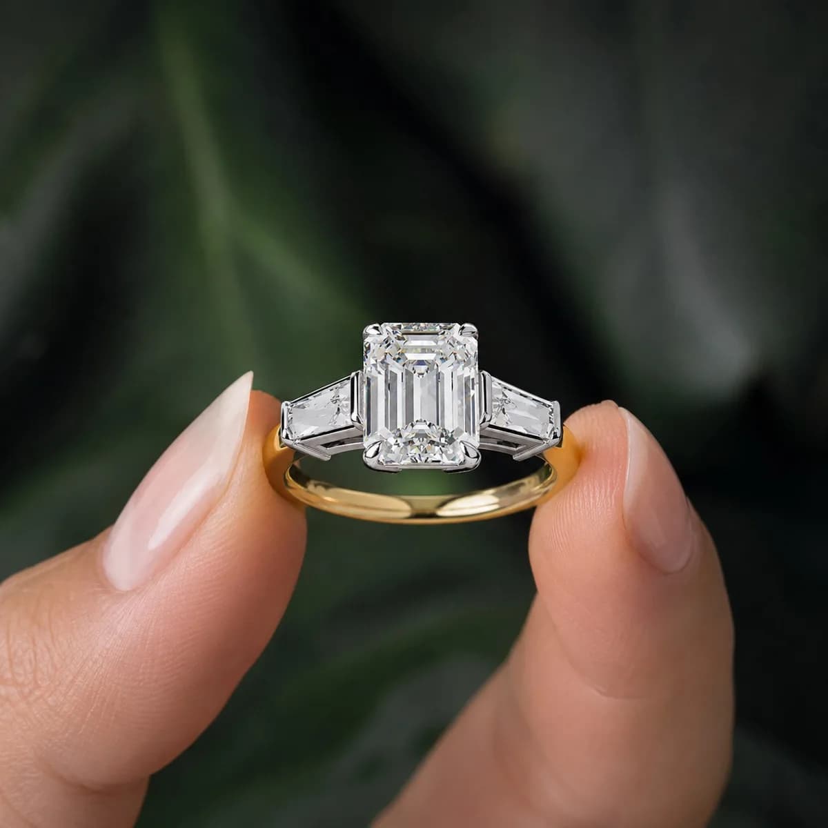 Emerald cut lab grown diamond three-stone engagement ring with baguettes in yellow gold held between fingertips