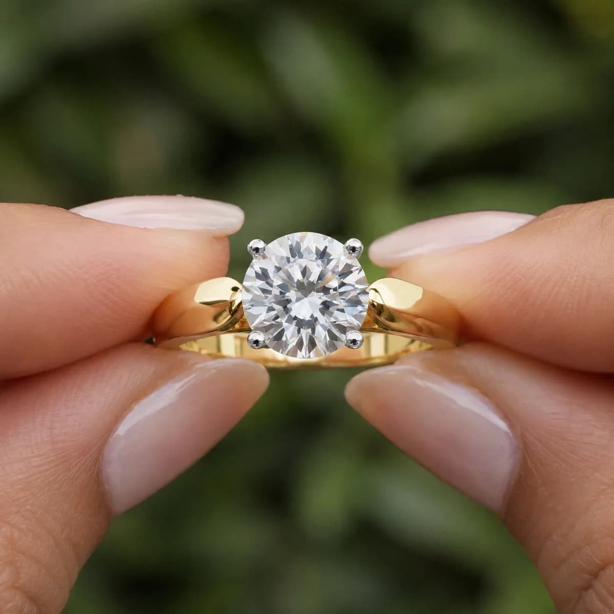 Round lab grown diamond four-claw cathedral solitaire engagement ring in yellow gold held between fingertips