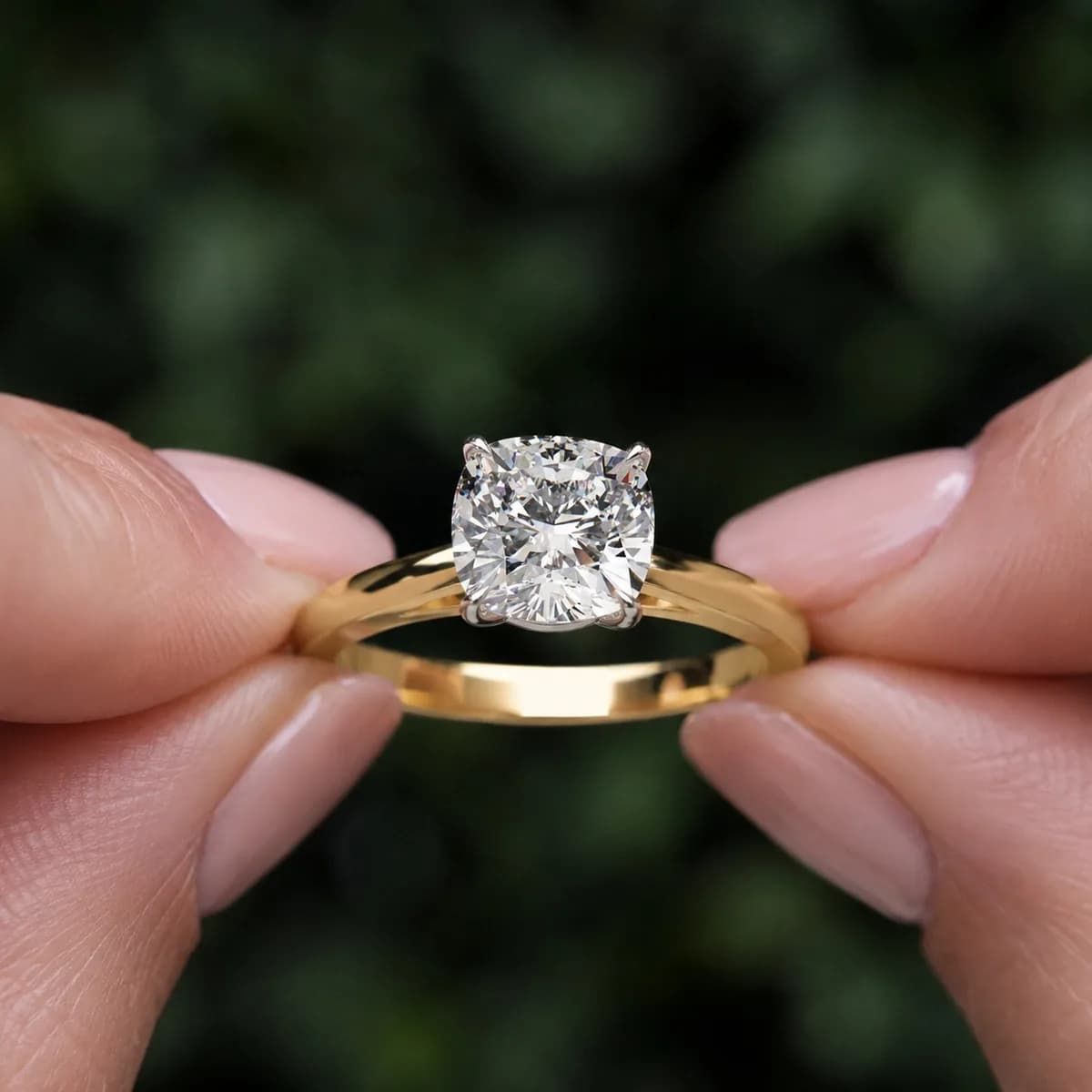 Cushion cut lab grown diamond cathedral solitaire engagement ring in yellow gold held between fingertips