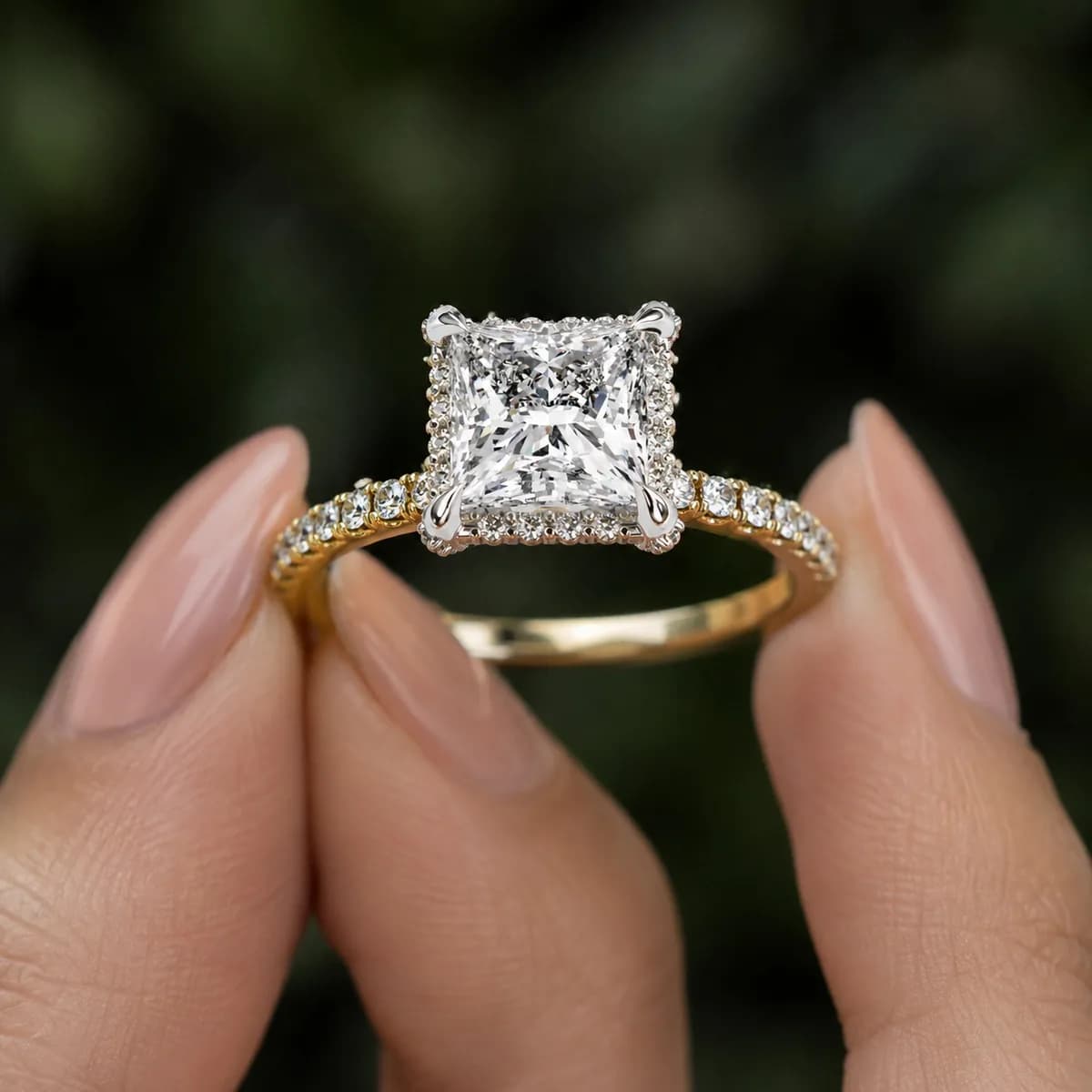 Princess cut lab grown diamond low halo engagement ring with pavé band in yellow gold held between fingertips